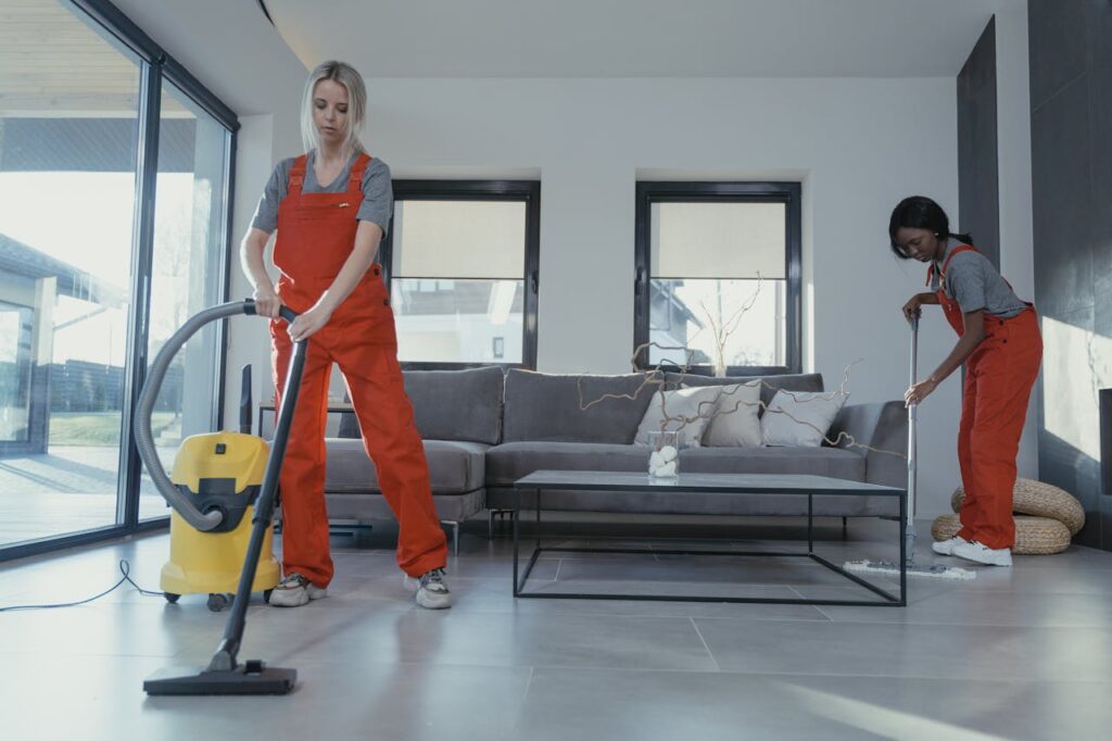 pexels-photo-6195277 Two women in red overalls vacuuming and sweeping a modern living room.